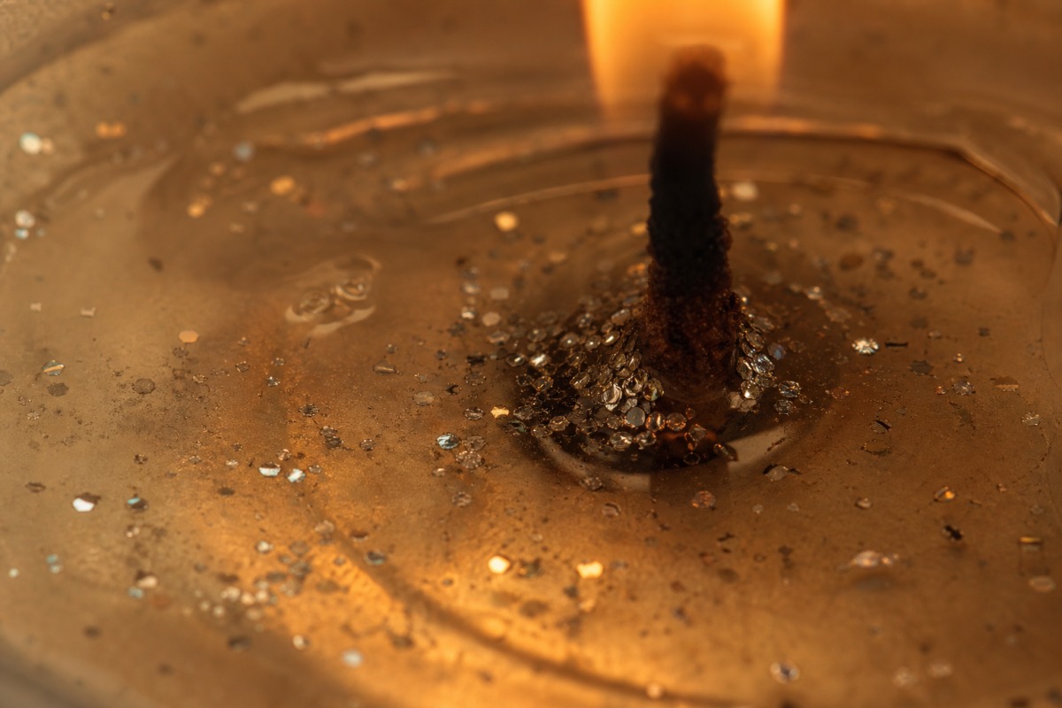 Macro shot of clear melted wax beside the wick of a burning candle, with tiny silver glitter particles sinking through it.
